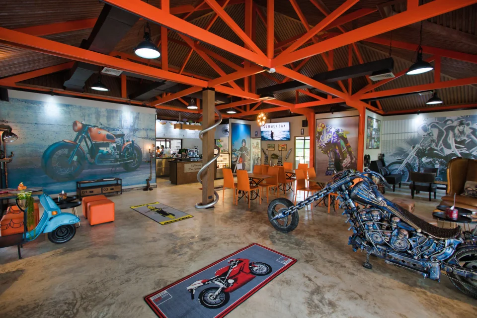 The dining room under orange steel roof trusses, with a steel chopper sculpture, a blue scooter and motorcycle murals