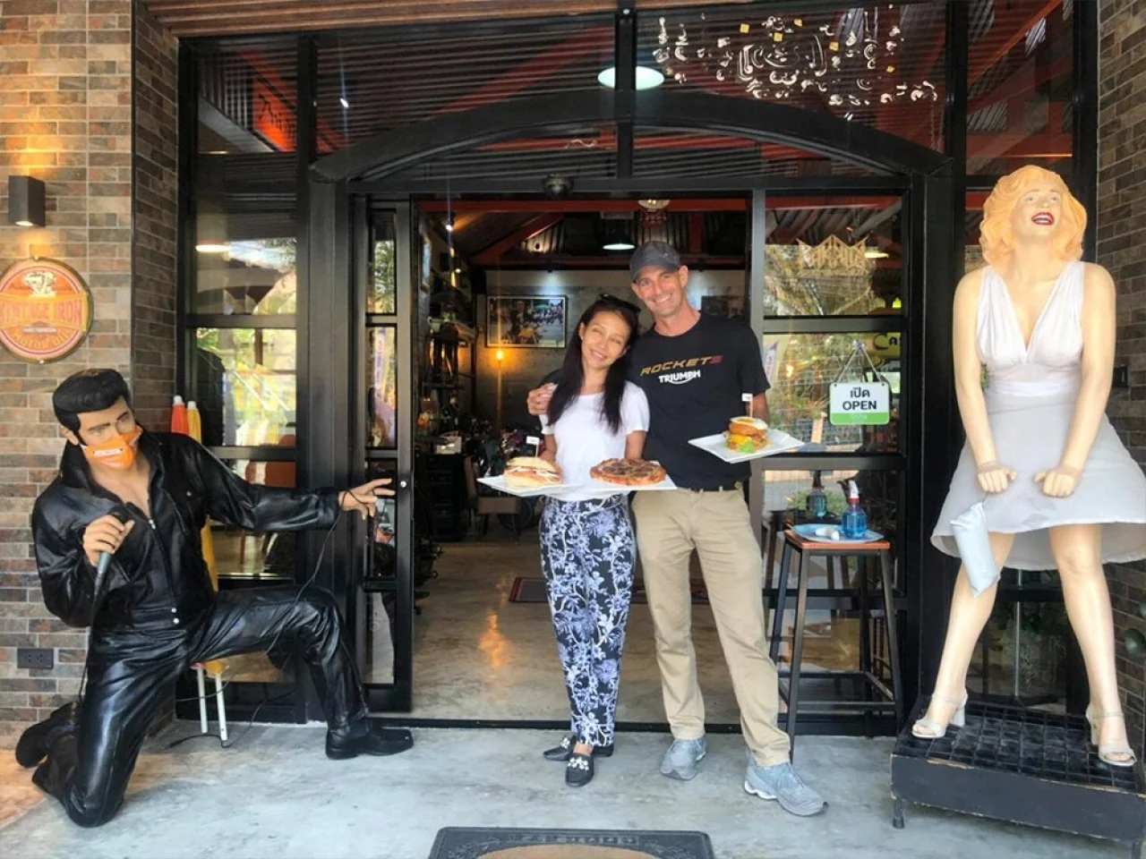 Achara and Martin at the front door with burgers and a pizza, between statues of Elvis and Marilyn Monroe