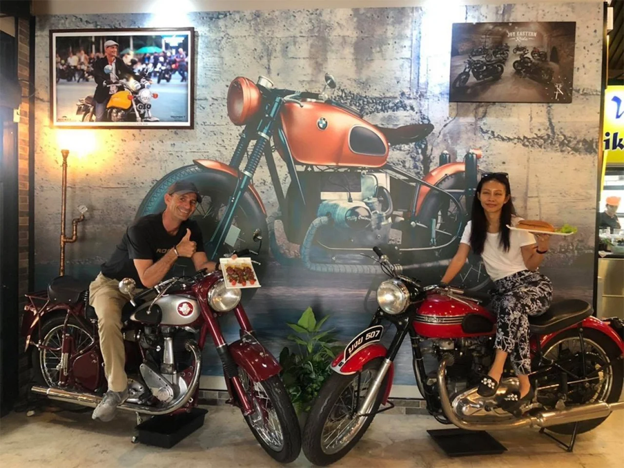 Achara and Martin with plates of food, sitting on two classic motorcycles in front of a mural of an orange BMW