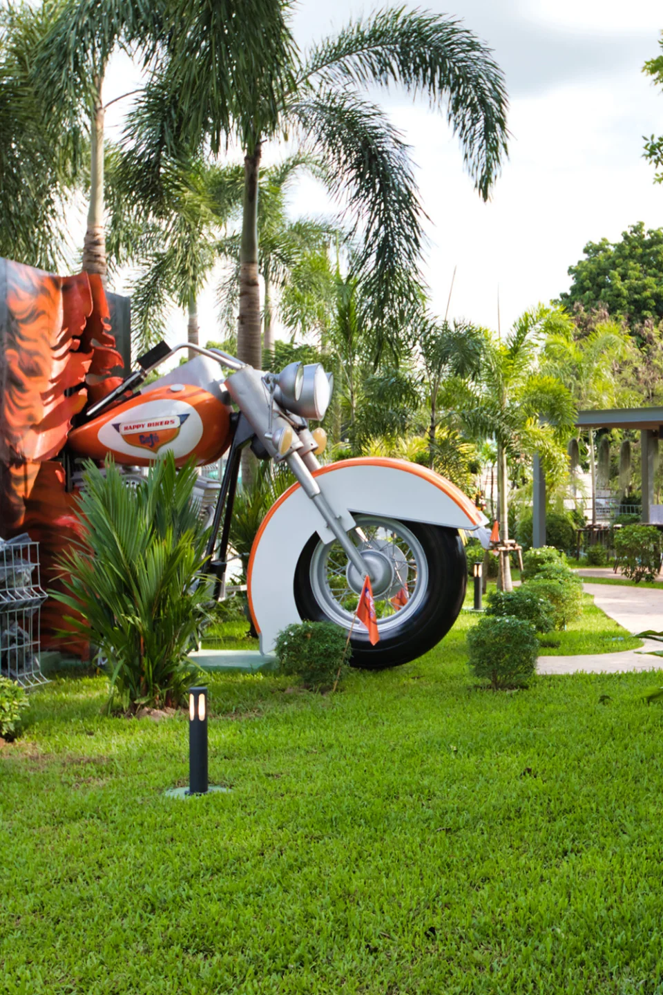 A giant orange and white motorcycle sculpture riding out of a flame-painted wall in the palm garden